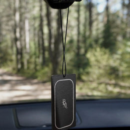 A Bombay Musk scented tag hanging from a car rear view mirror on a forest road