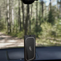 A Bombay Musk scented tag hanging from a car rear view mirror on a forest road